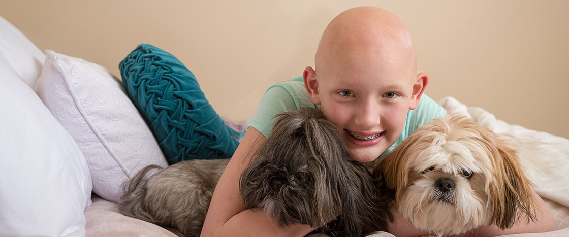 A girl with no hair smiling and hugging two cute dogs