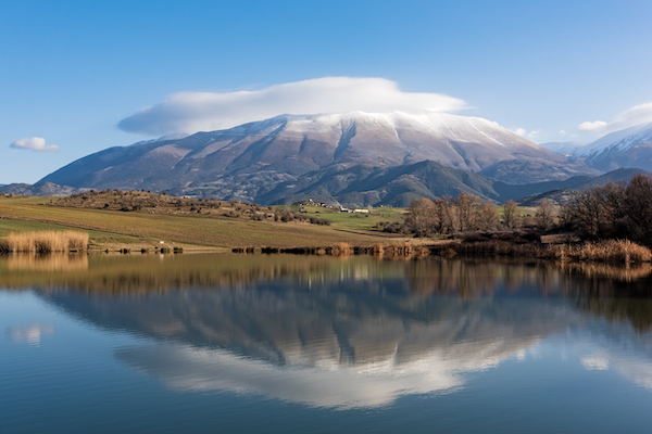 A mountain with snow capped top and clouds floating behind it 